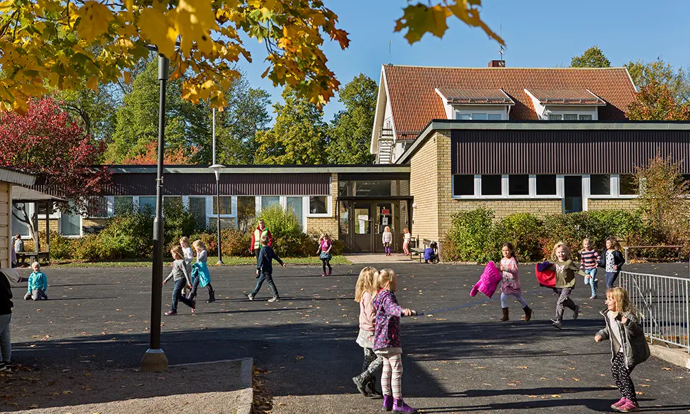 Foto av Grillbyskolan. Det är en gul tegelbyggnad. I förgrunden syns elever på skolgården.