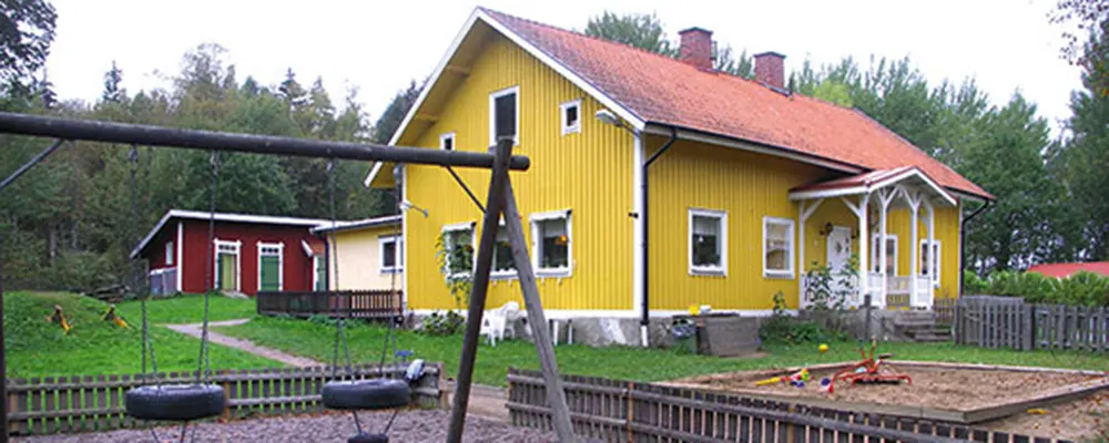 Foto av Palettens förskola i Ekolsund. Det är ett gul trähus med vita knutar. I förgrunden syns lekutrustning. 